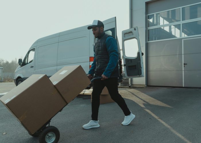 Courier pushing a trolley with boxes near a van at a warehouse, showcasing delivery services.