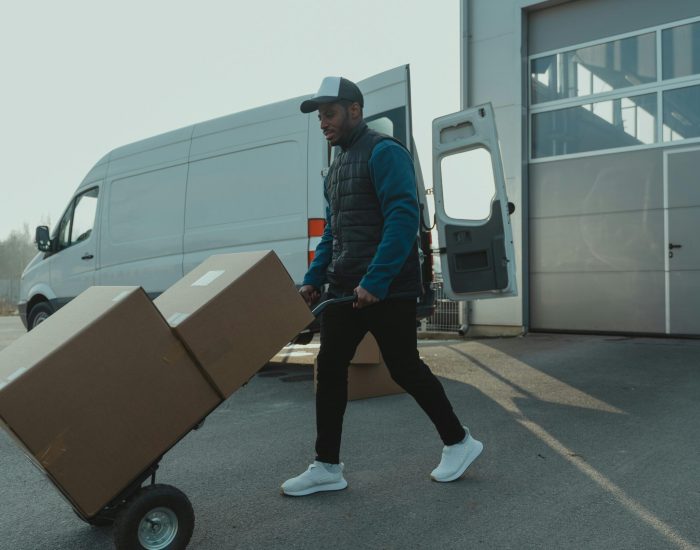 Courier pushing a trolley with boxes near a van at a warehouse, showcasing delivery services.