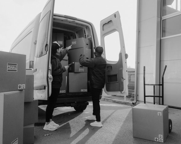 Two couriers loading cardboard boxes into a delivery van outdoors.